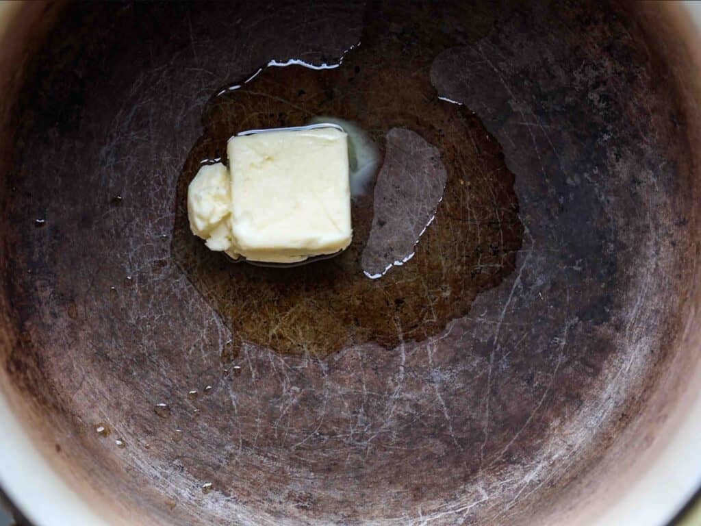A cube of vegan butter melting in a large, well-used pan, with liquid butter starting to spread around it.